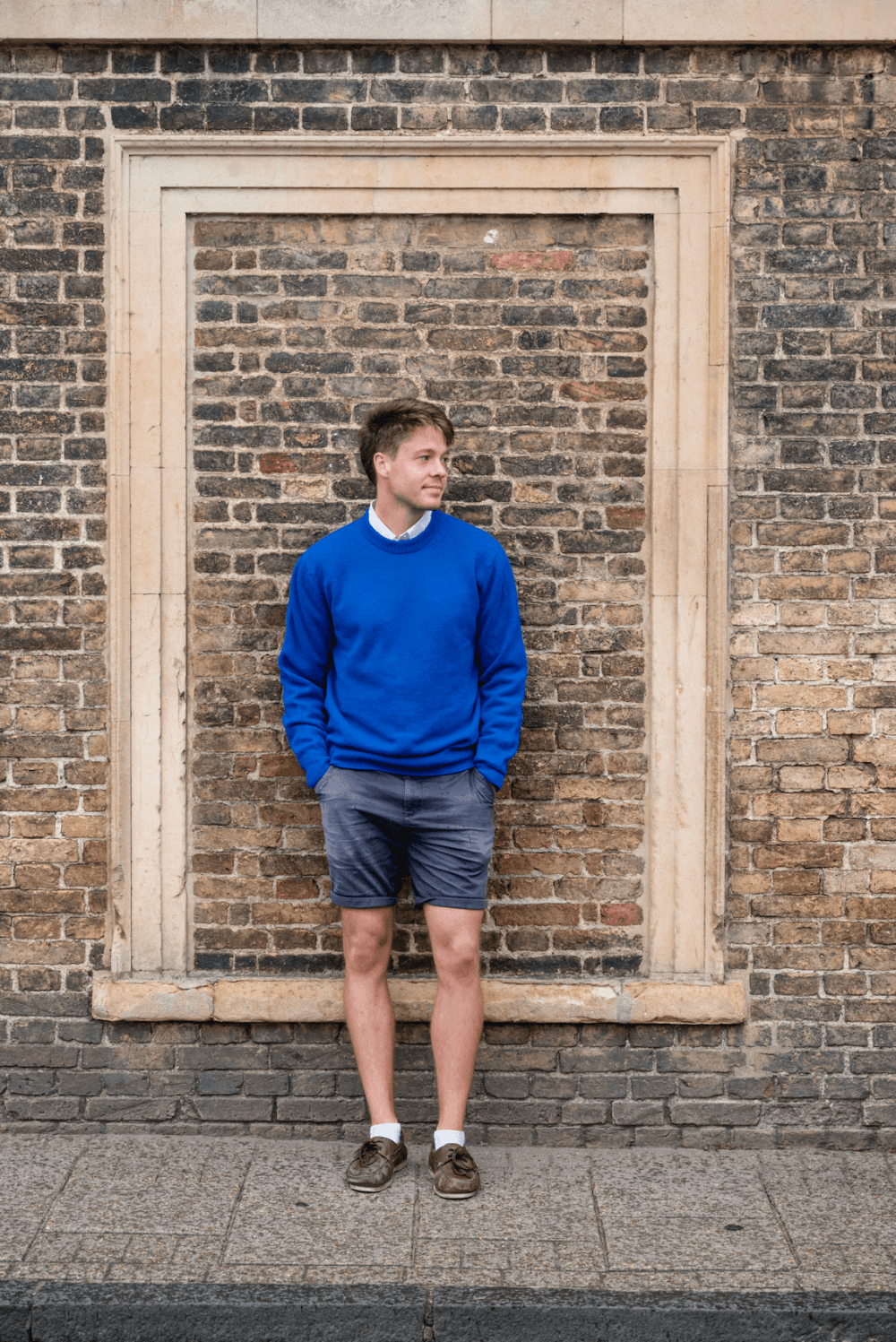 Man wearing a blue sweater and shorts standing in front of a brick wall 