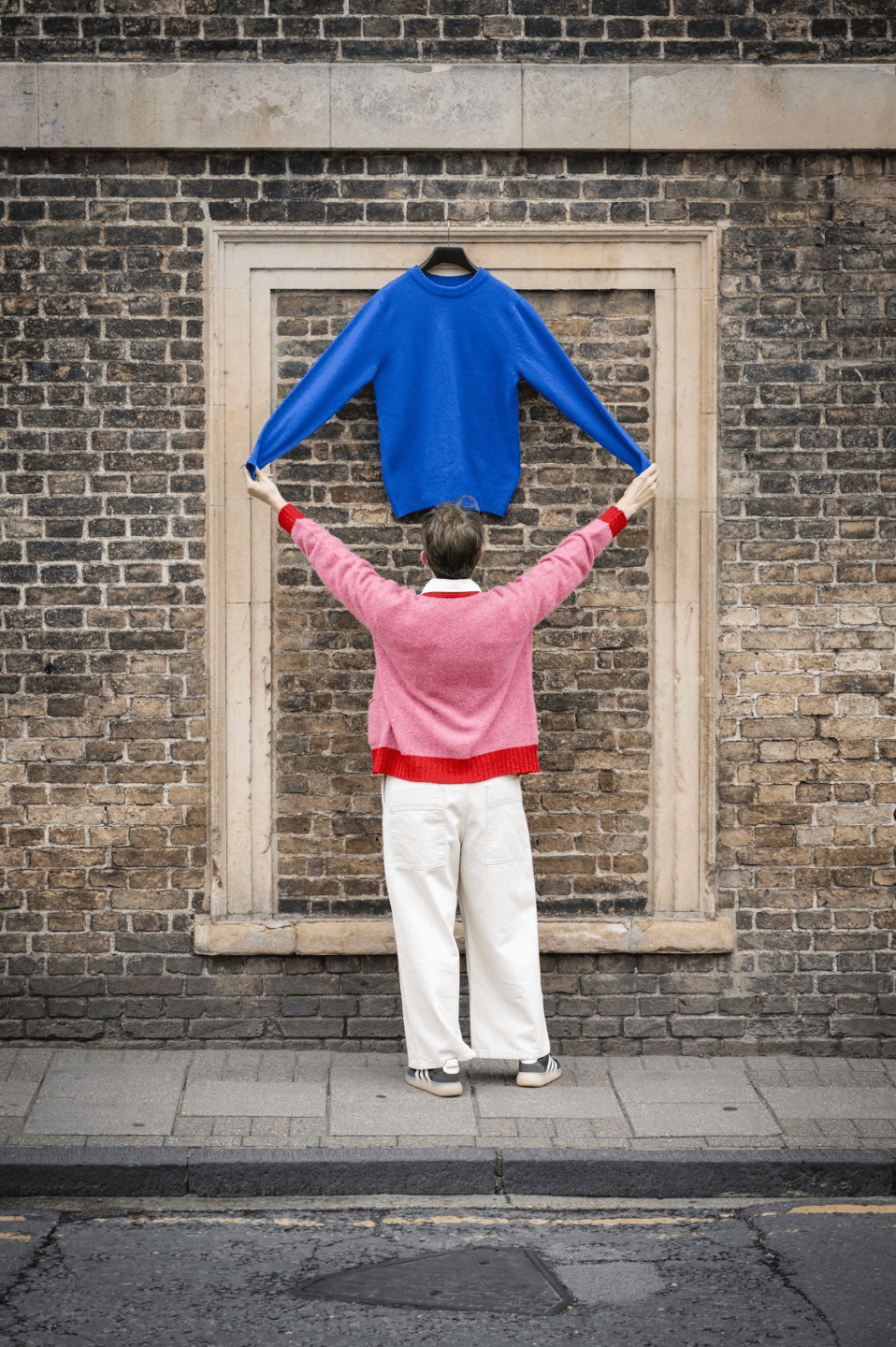 Blue crew neck jumper hanging in front of a brick wall with woman in pink cardigan